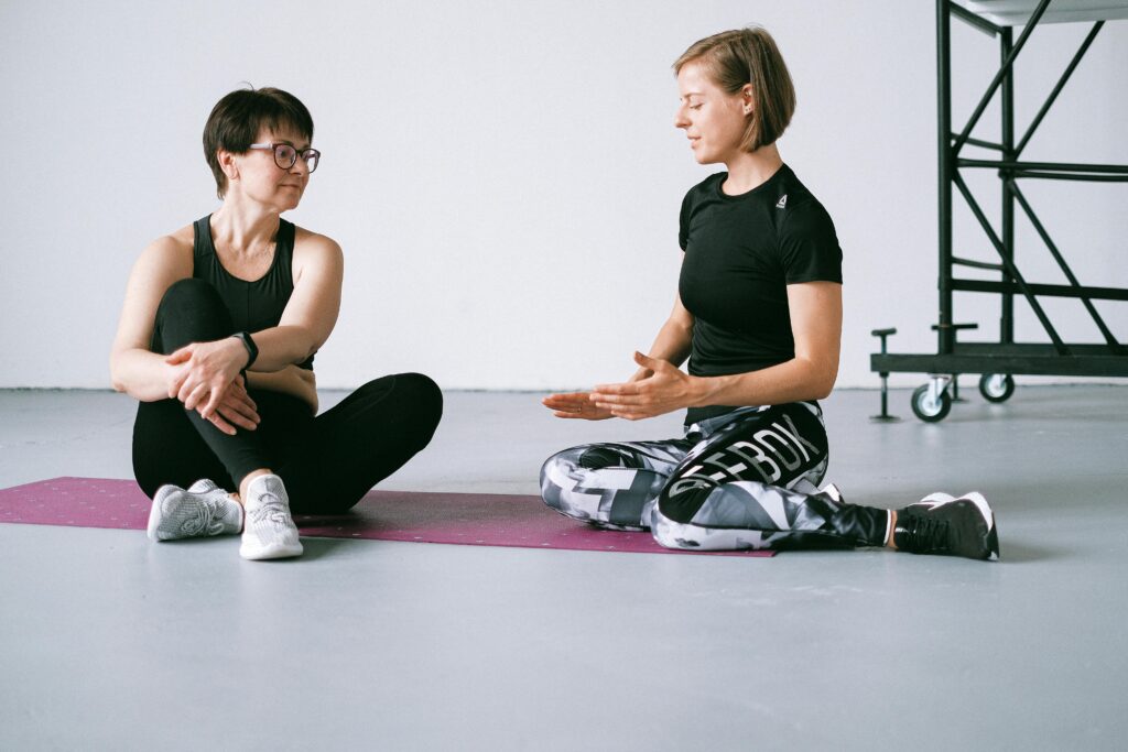 Two women in active wear sitting on yoga mats discussing exercise in a gym setting.