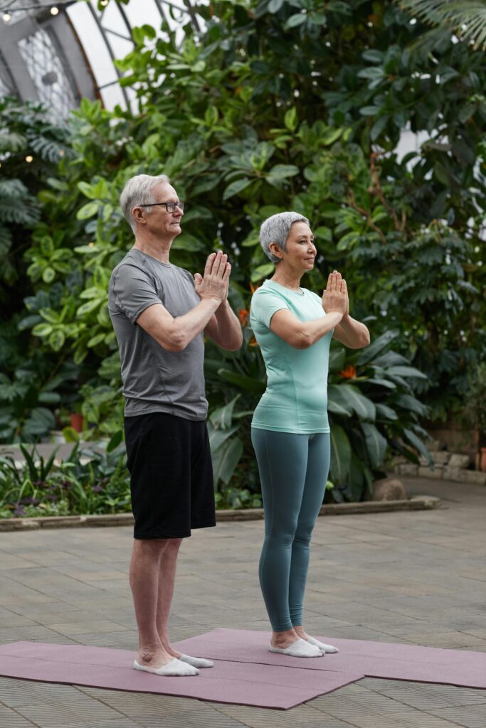 Senior couple practicing yoga poses together in a lush greenhouse setting.