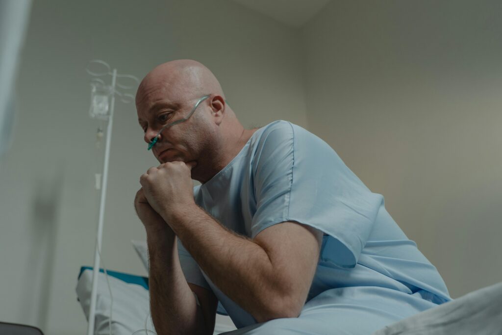 Bald man in hospital gown sits pensively with oxygen tube in healthcare setting.
