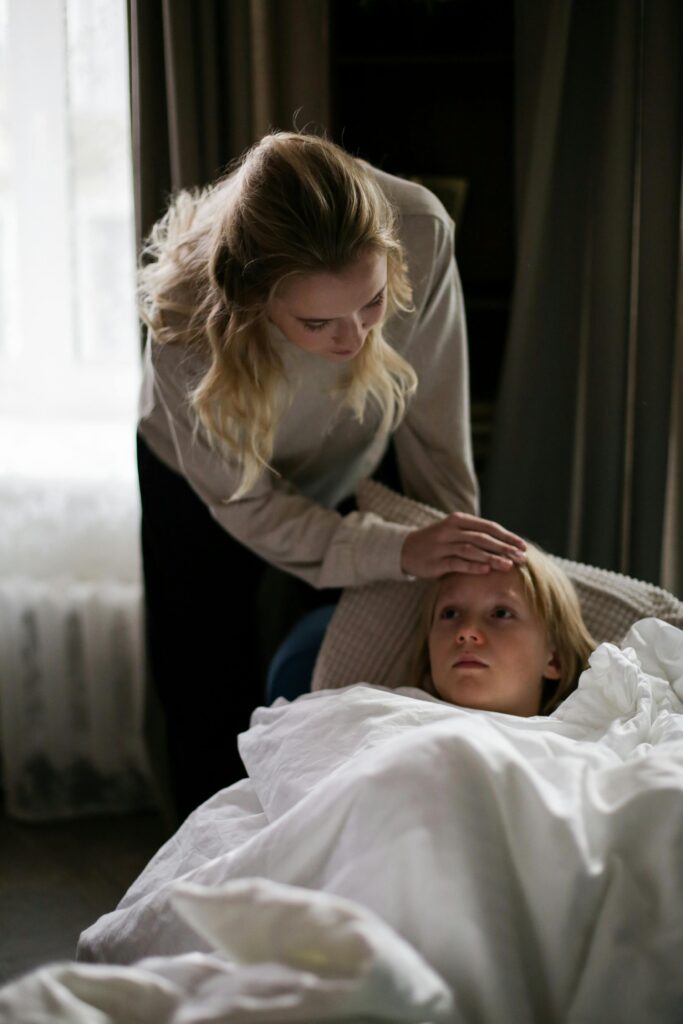 A concerned mother soothing her sick child lying on a couch under a white blanket indoors.