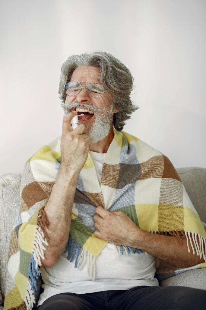 Elderly man using throat spray, wrapped in a cozy checkered blanket, healthcare theme.