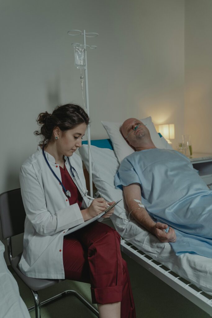 A doctor sits beside a patient in a hospital room, taking notes.