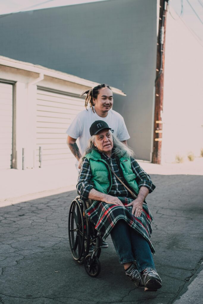 A helpful young adult pushes a senior in a wheelchair on a sunny day, promoting kindness.