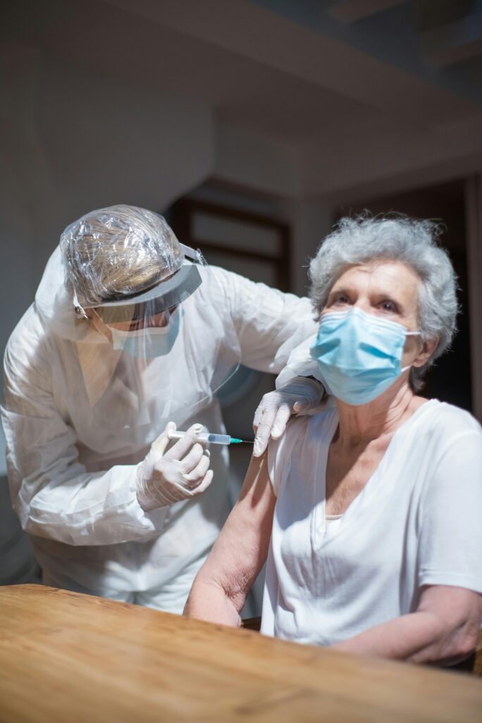 Elderly woman receiving a vaccine from a healthcare worker in protective gear indoors.